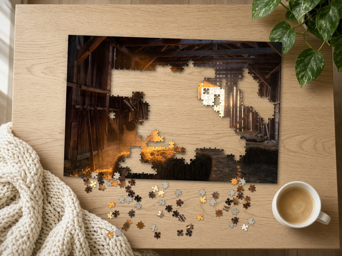 The Barn That Held the Day 1014 piece puzzle half assembled on a light oak table, loose pieces scattered beside a chunky knit throw, a latte and a trailing plant