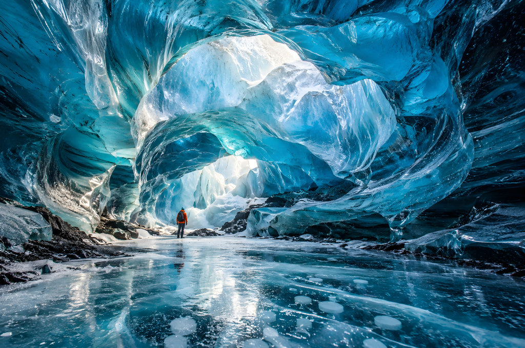 The Cave Where the Ice Glows Blue art puzzle: a glowing blue ice cave arching over a frozen floor, a small explorer in an orange jacket standing in the light at the far end