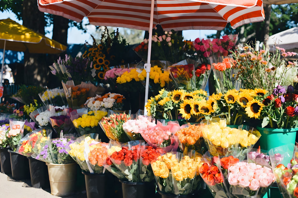 The Flower Stall on Market Morning art puzzle: a sunny outdoor flower stall under a red and white striped umbrella, buckets of sunflowers, roses and tulips in wrapped bunches