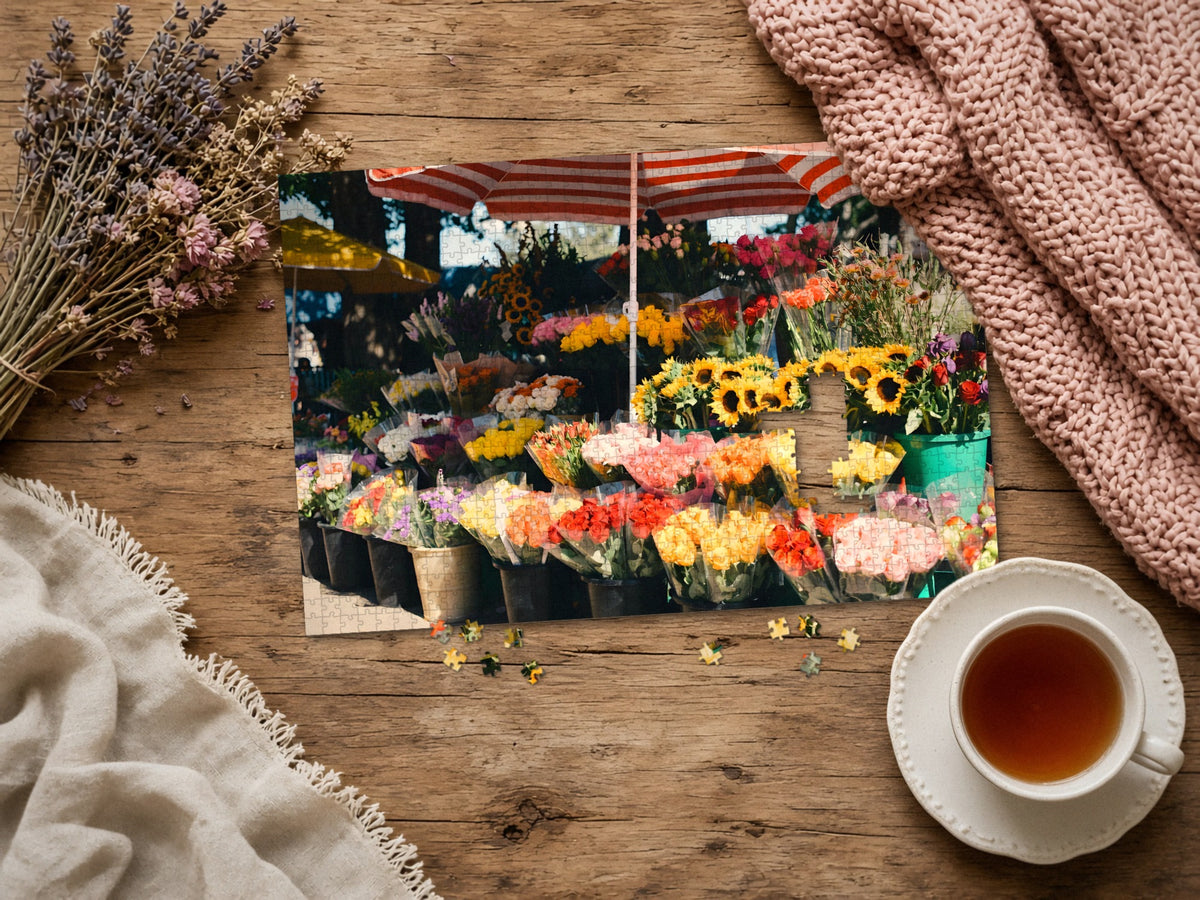 The Flower Stall on Market Morning 1014 piece puzzle assembled on a rustic wood table with dried lavender, a pink knit blanket, a linen throw and a cup of tea, a few loose pieces nearby