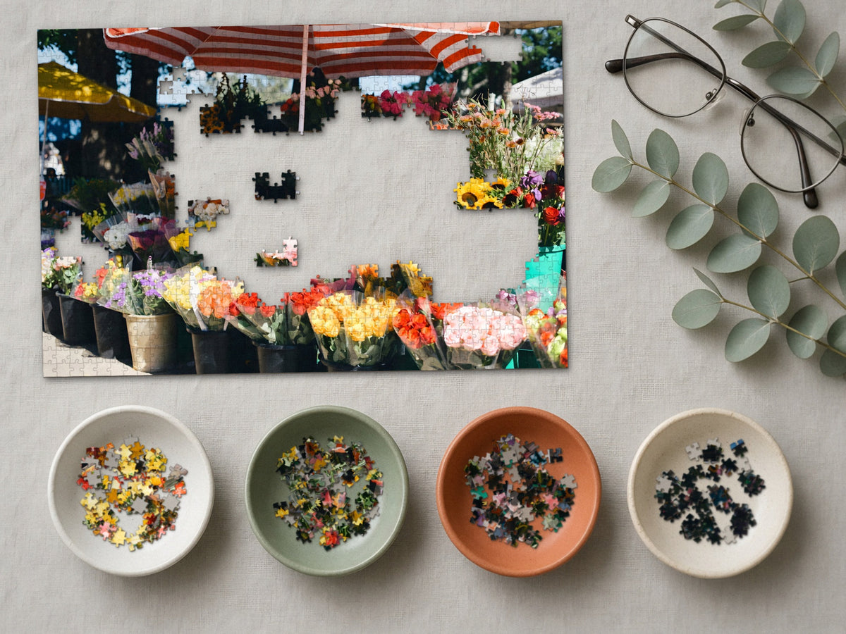 The Flower Stall on Market Morning 1014 piece puzzle partly assembled on a linen cloth, loose pieces sorted into four small ceramic bowls beside reading glasses and eucalyptus
