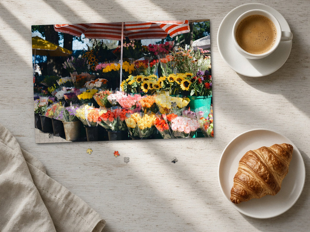 The Flower Stall on Market Morning 1014 piece puzzle laid flat on a pale wood table in morning sun beside a cappuccino and a croissant