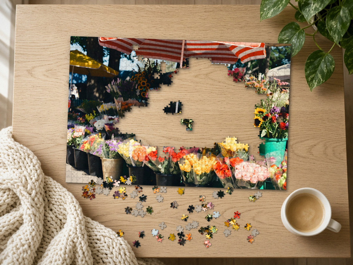 The Flower Stall on Market Morning 1014 piece puzzle half assembled on a light oak table, loose pieces scattered beside a chunky knit throw, a latte and a trailing plant