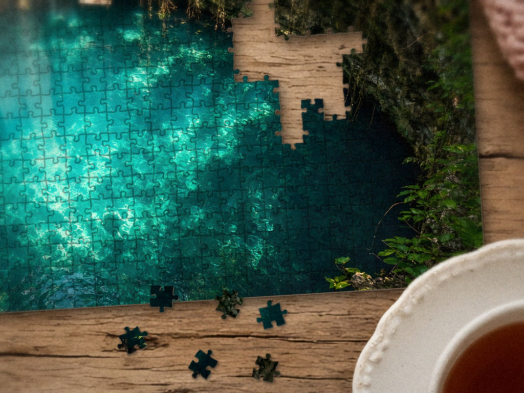 Close-up of the The Pool the Jungle Kept Secret puzzle on a wood table showing the teal water glowing under the light shafts, a few pieces still missing, next to a teacup