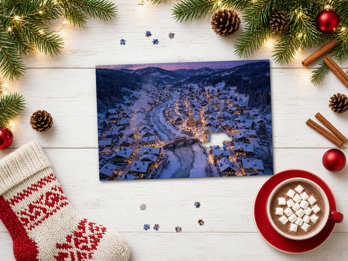 The Village the Snow Tucked In 1014 piece puzzle on a white wood table with pine branches, fairy lights, pinecones, red ornaments, cinnamon sticks, a knit stocking and a mug of cocoa with marshmallows