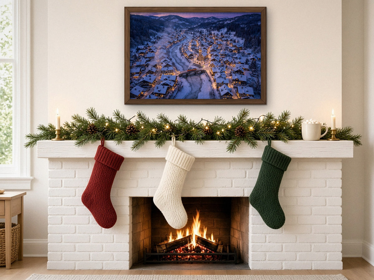The Village the Snow Tucked In 1014 piece puzzle assembled, framed in dark wood above a white brick fireplace with a pine garland and red, cream and green stockings