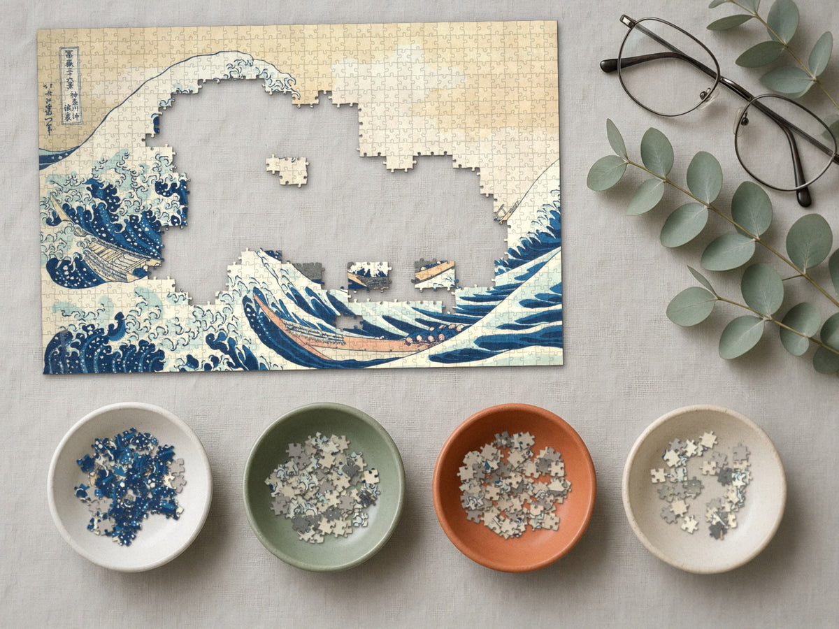 The Wave Before the Mountain 1014 piece puzzle partly assembled on a linen cloth, loose pieces sorted into four small ceramic bowls beside reading glasses and eucalyptus
