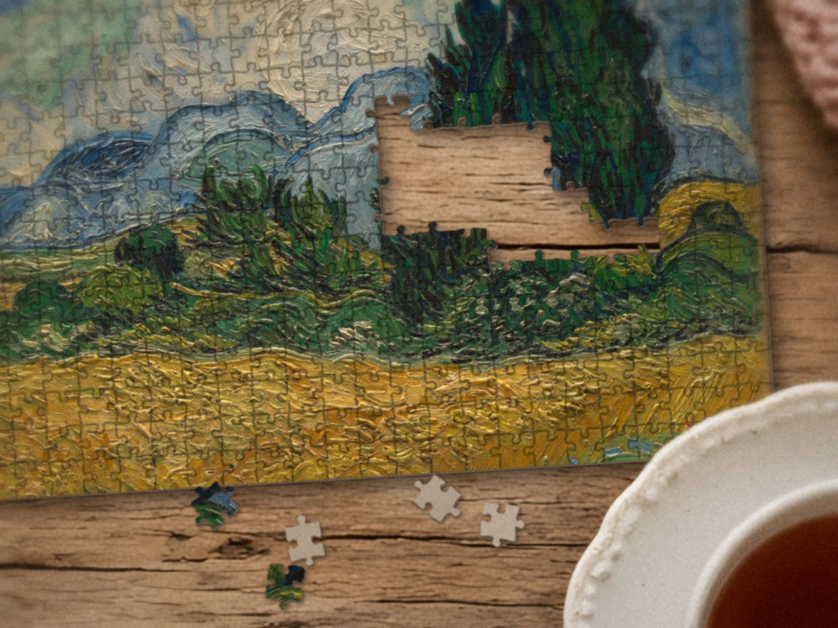 Close-up of the Wind Over the Wheat puzzle on a wood table showing the cypresses and wheat, a few pieces still missing, next to a teacup
